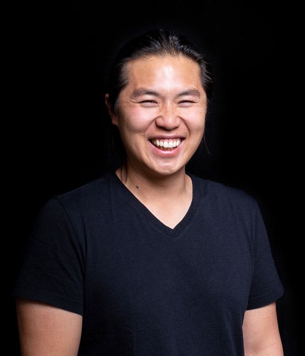 An asian man with pulled back black hair smiling broadly while wearing a black shirt, on a black background.