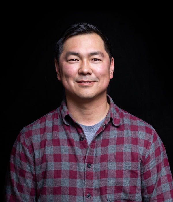 A smiling asian man with short black hair, wearing a red and blue flannel shirt on a black background.