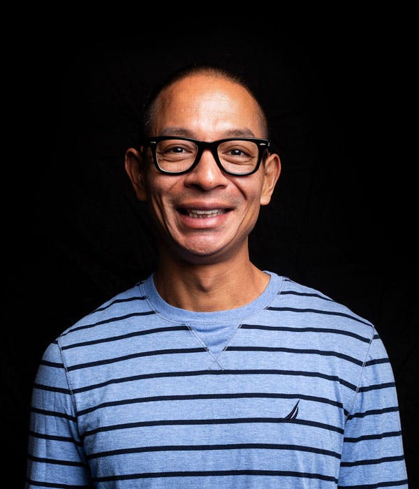 A smiling bald asian man with black glasses wearing a striped blue shirt on a black background.