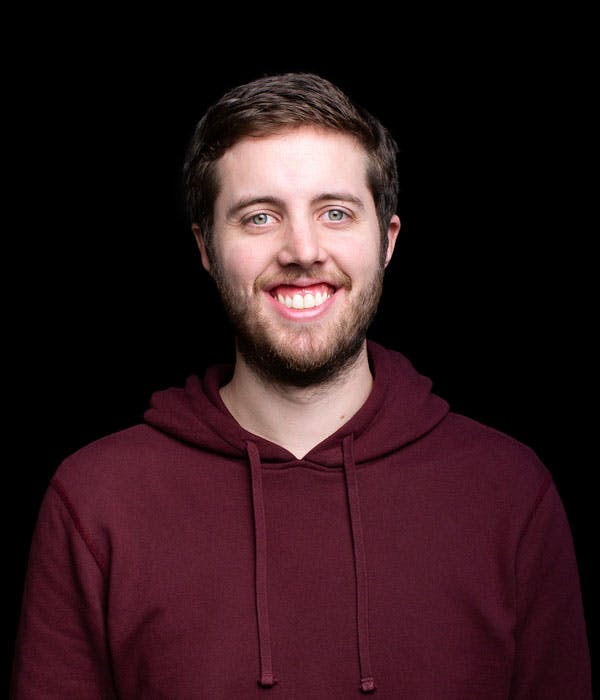 A white man with brown hair smiling broadly, wearing a wine colored hoodie, on a black backdrop.