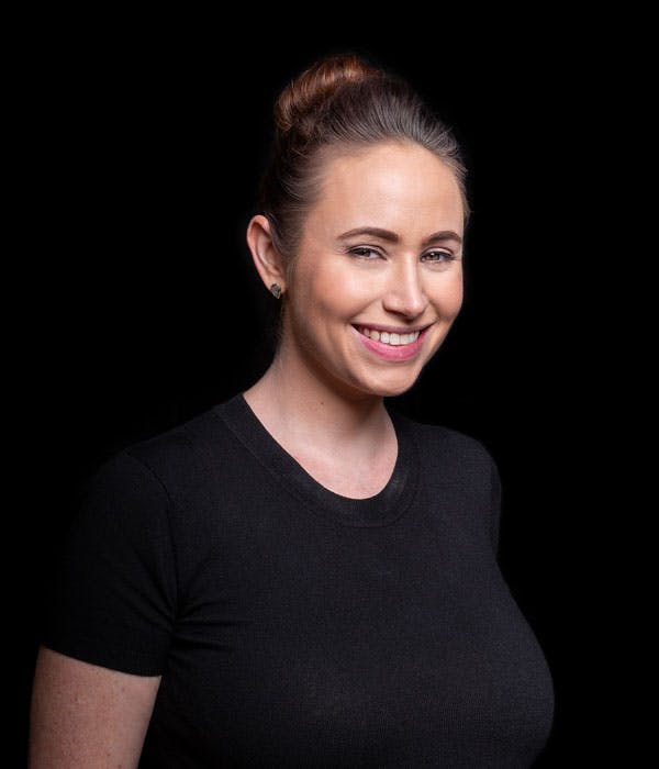 A smiling white woman with brown hair in an updo, wearing a black shirt, on a black background.