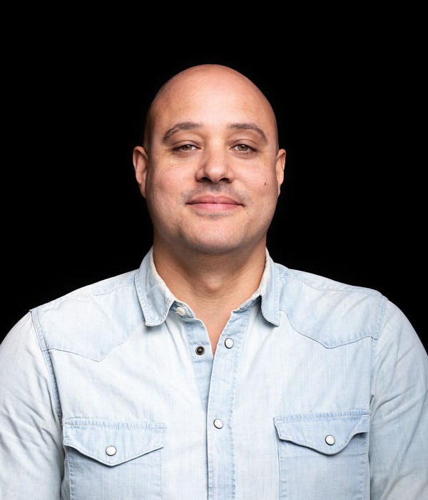 A smiling bald man with a blue button up shirt on a black background.