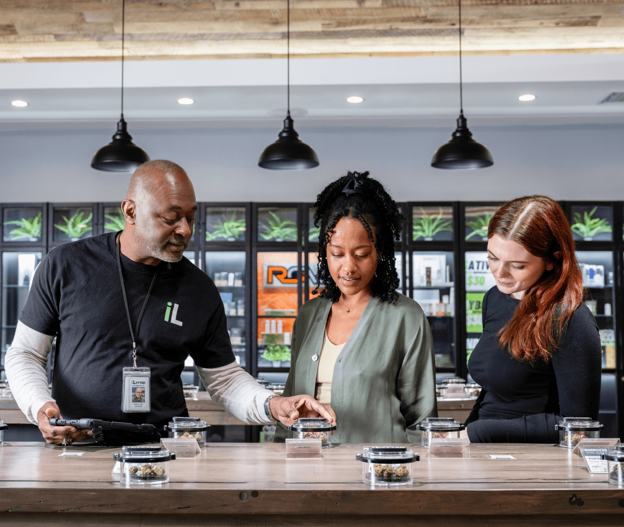 A dispensary employee shows two customers cannabis flower displayed in clear containers on a countertop. The employee holds a tablet while guiding the customers, who are closely examining the products. Behind them, shelves are stocked with cannabis jars and signage, creating a bright, modern retail environment.
