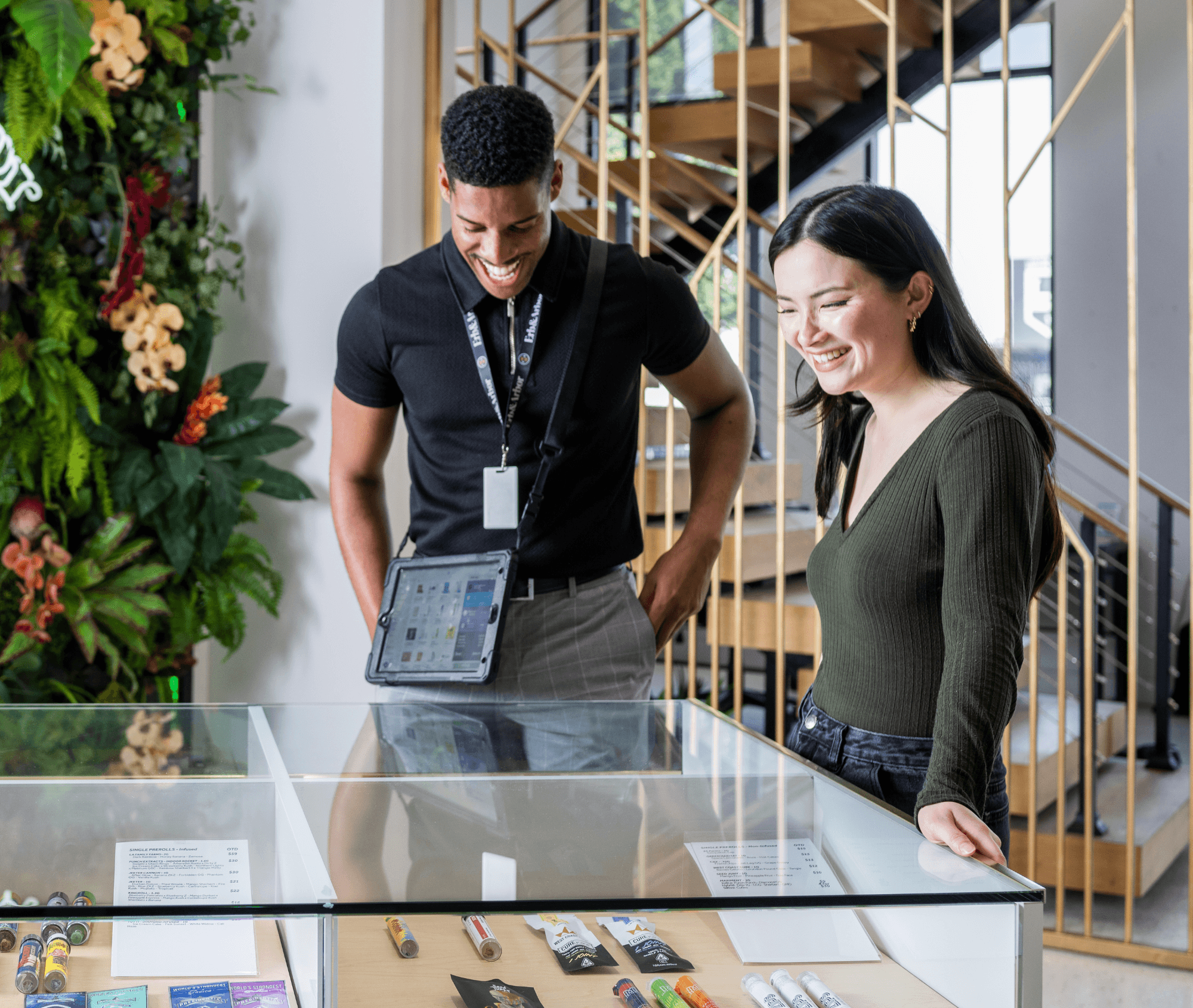 A smiling budtender and customer stand at a glass display case inside a bright, modern dispensary. The budtender wears a Meadow tablet around his neck, showing the POS interface. Behind them is a staircase and a lush vertical plant wall, reflecting a clean, welcoming retail environment.