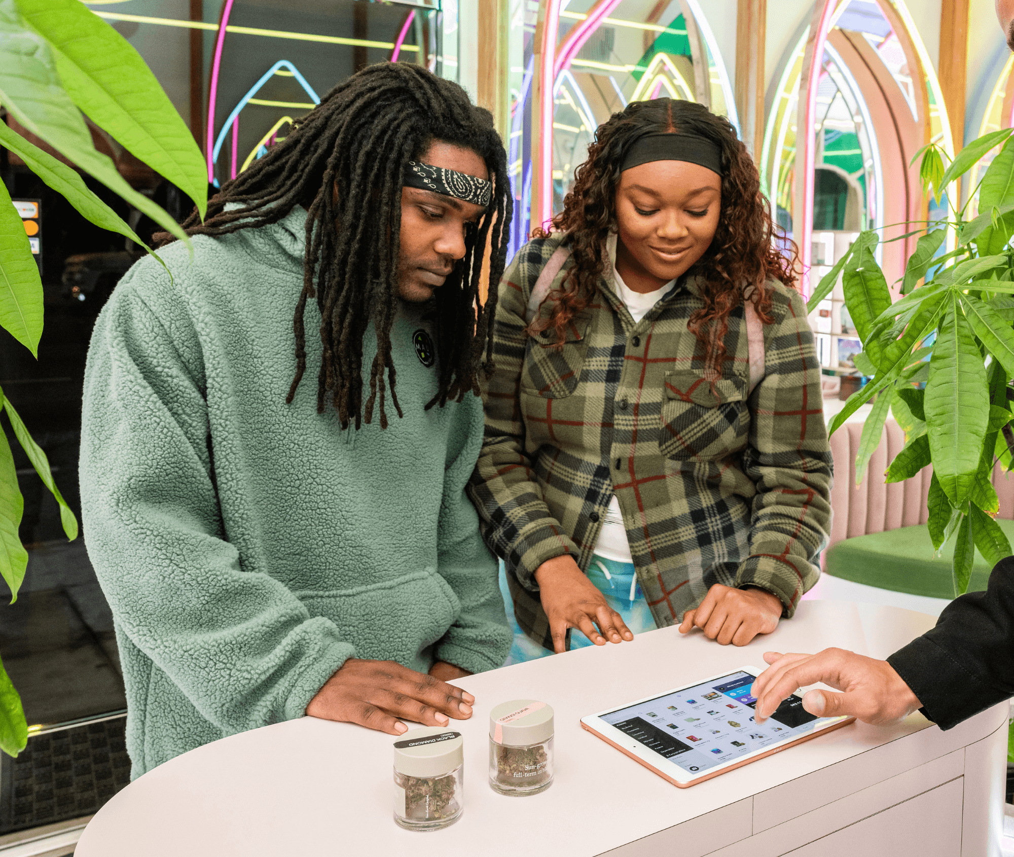 Two customers examine cannabis flower jars at a dispensary counter as a staff member uses a Meadow-powered tablet to guide them through menu options. The setting highlights interactive, compliant retail workflows that support customer engagement.