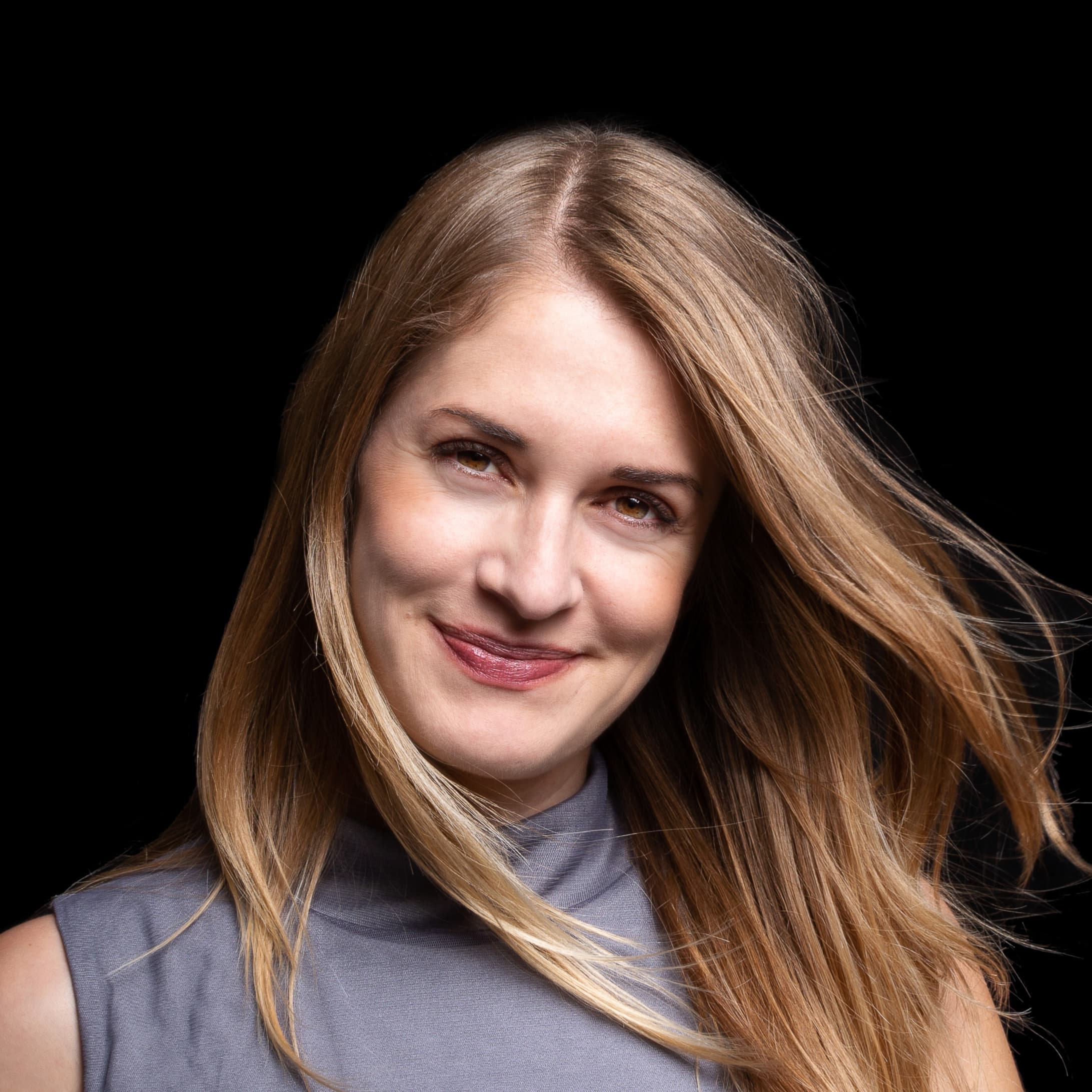 A smiling white woman with long, flowing blonde hair wearing a sleeveless gray top.