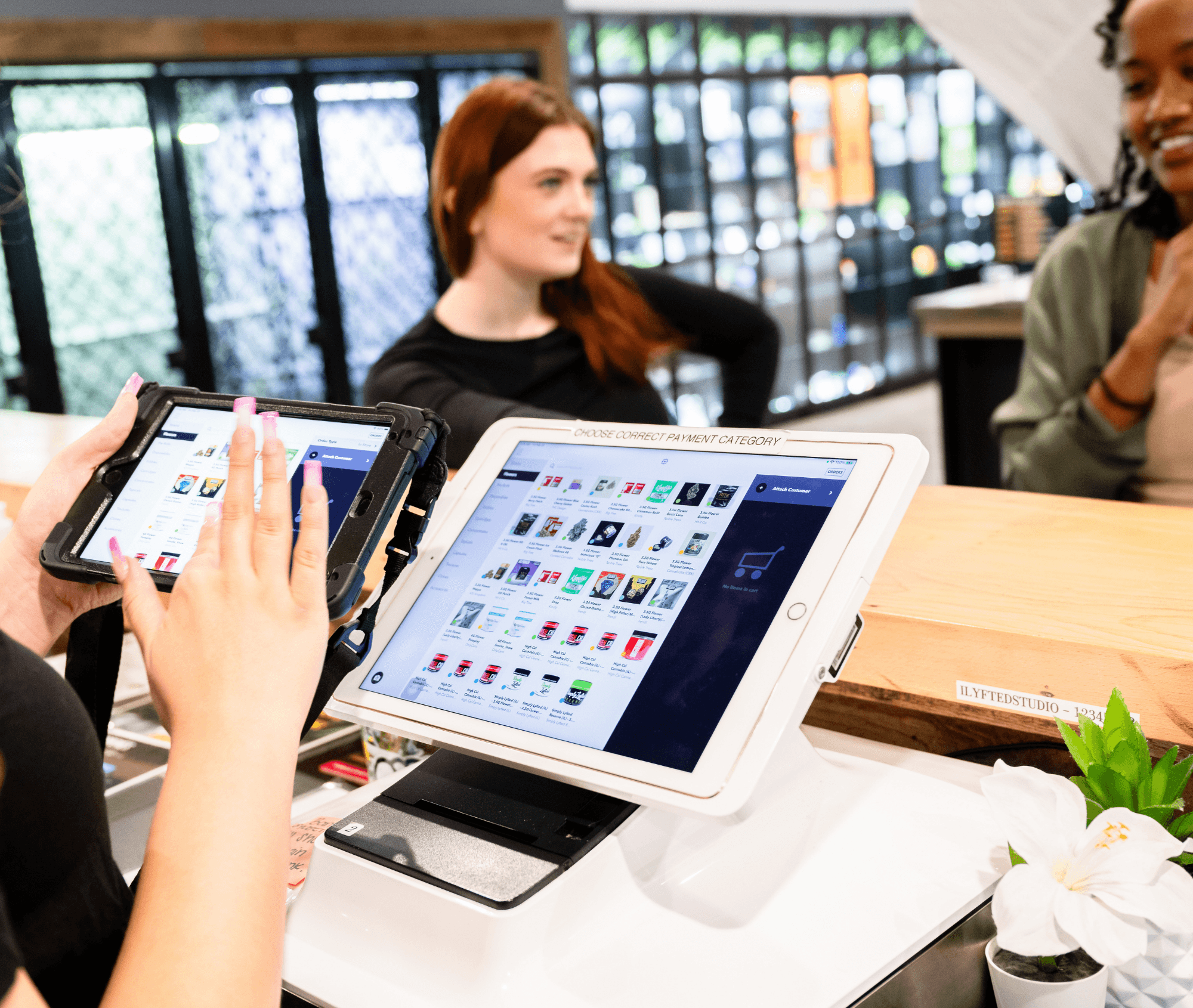 Close-up of a dispensary point-of-sale system showing cannabis product menus on a tablet and handheld device. In the background, staff engage with customers at the counter, highlighting a professional, modern retail environment designed for efficient sales and customer service.
