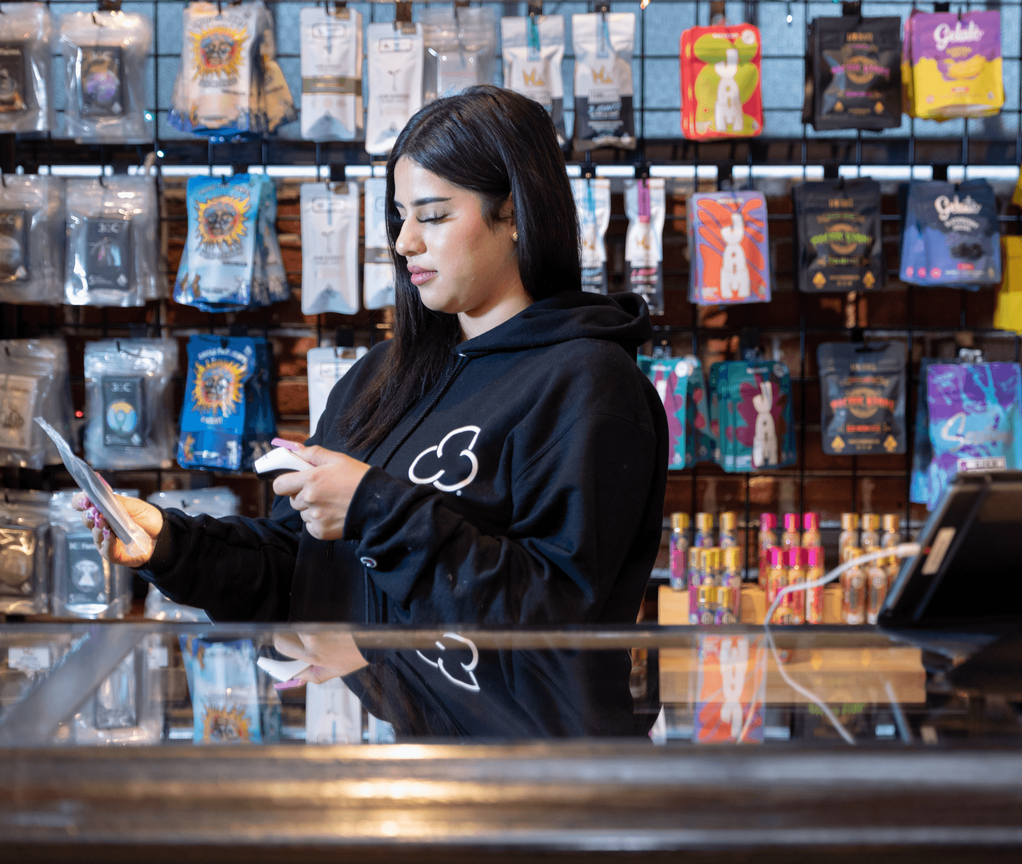 A budtender in a black hoodie scans cannabis product packaging behind a glass counter, with a colorful, wall-mounted display of sealed cannabis goods in the background. The setting is a well-lit, organized dispensary.