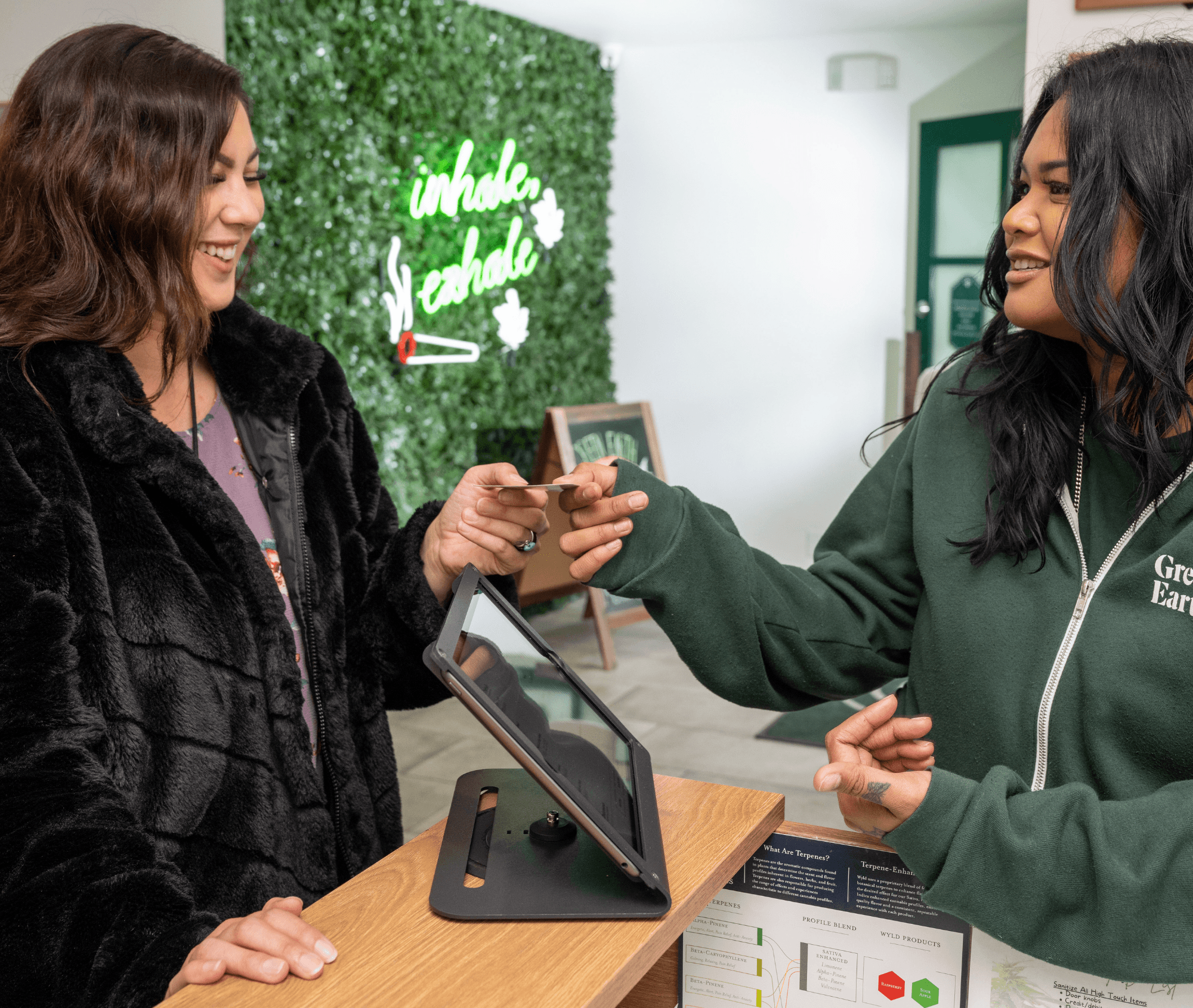 A budtender hands a customer their debit card during check-out at a dispensary register powered by Meadow. The setting features a “inhale, exhale” neon sign on a greenery wall, creating a relaxed and friendly atmosphere at check-out